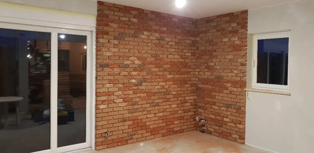 Wnętrze z fragmentem ściany wykończonym dekoracyjną cegłą w odcieniach czerwieni i brązu, widoczne białe okno i przesuwne drzwi balkonowe z żółtą taśmą malarską na górze, surowy stan wykończenia...