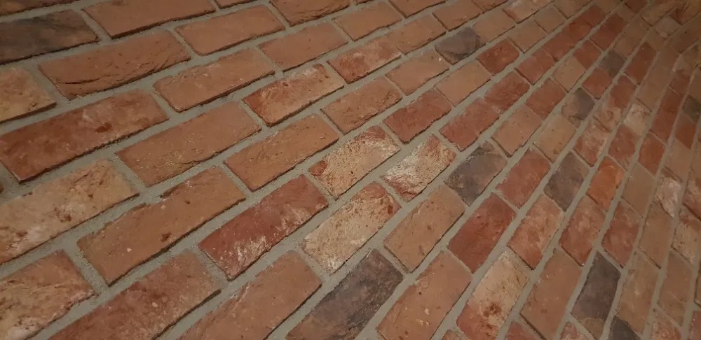 Ukośny widok na ścianę wyłożoną cegłami w odcieniach czerwieni i brązu z szarą fugą, tworzącą rustykalny wzór.