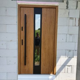 Stolar Agnieszka Pikulik - Zewnętrzne drzwi wejściowe z drewnopodobnym wykończeniem i pionowym przeszkleniem, zamontowane w surowym stanie budynku z białych bloczków, z widocznym odbiciem fotografa w szybie.