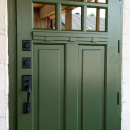 Zielone drzwi zewnętrzne z szybami w górnej części, czarnymi okuciami i widoczną konstrukcją budynku w tle przez szyby.