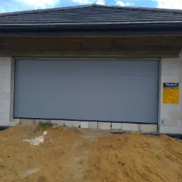 Szara brama segmentowa garażowa w surowym stanie, widoczna tablica informacyjna producenta Abakon, teren wokół w trakcie prac ziemnych, ściany z bloczków betonowych.