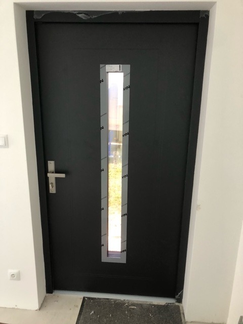 Ciemnoszare drzwi wejściowe z wąskim, pionowym przeszkleniem, częściowo zabezpieczonym folią ochronną, zamontowane w jasnym wnętrzu.