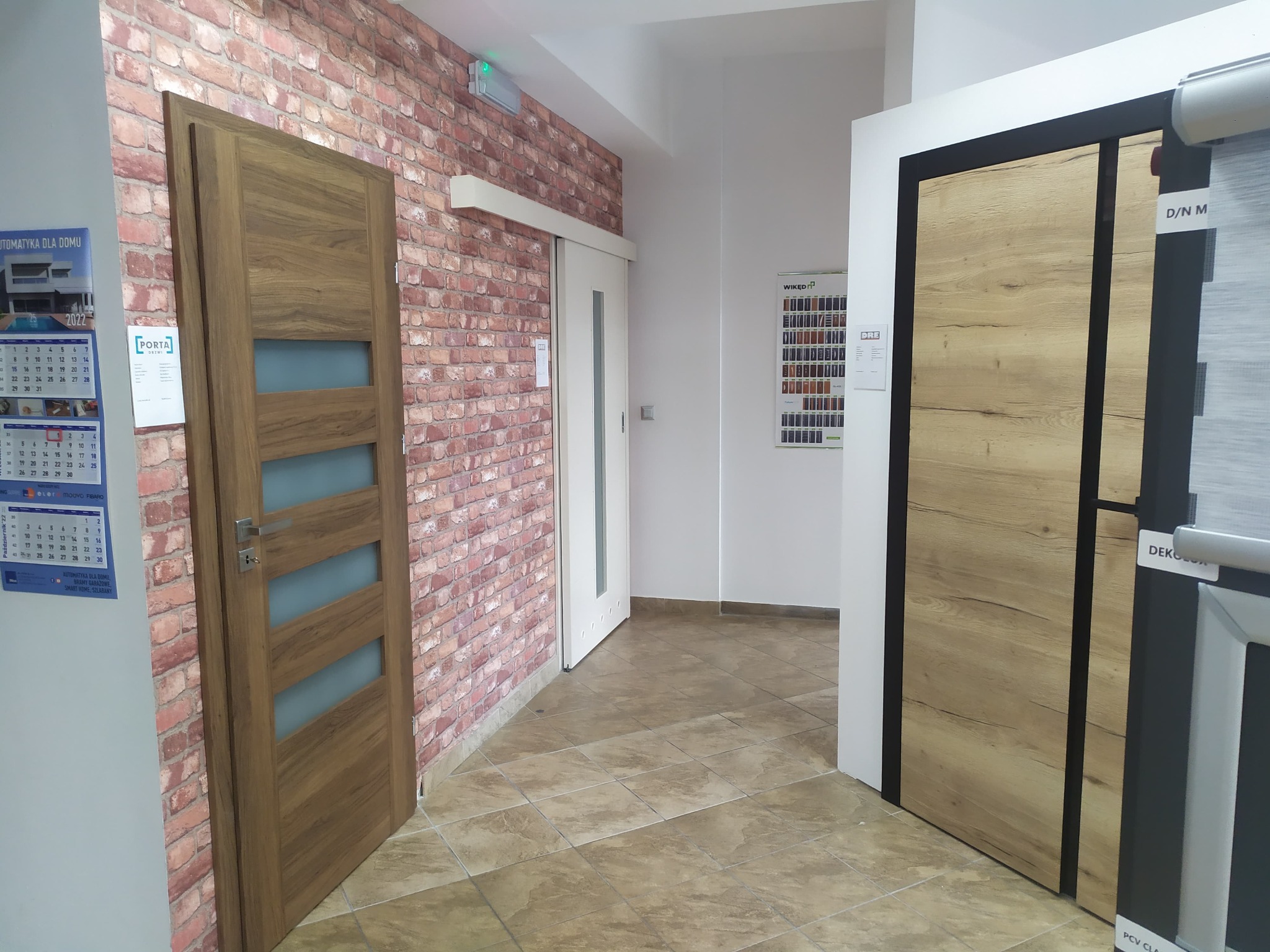 Wystawa w salonie z ekspozycją różnych modeli drzwi wewnętrznych: drewniane z przeszkleniami na tle ceglanej ściany, przesuwne białe oraz z okleiną drewnopodobną z czarnymi obramowaniami,...