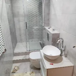 Szara łazienka z kabiną prysznicową z wzorem, umywalką nablatową na białej szafce i wiszącą toaletą. Na podłodze leży dywanik z dziecięcym motywem.