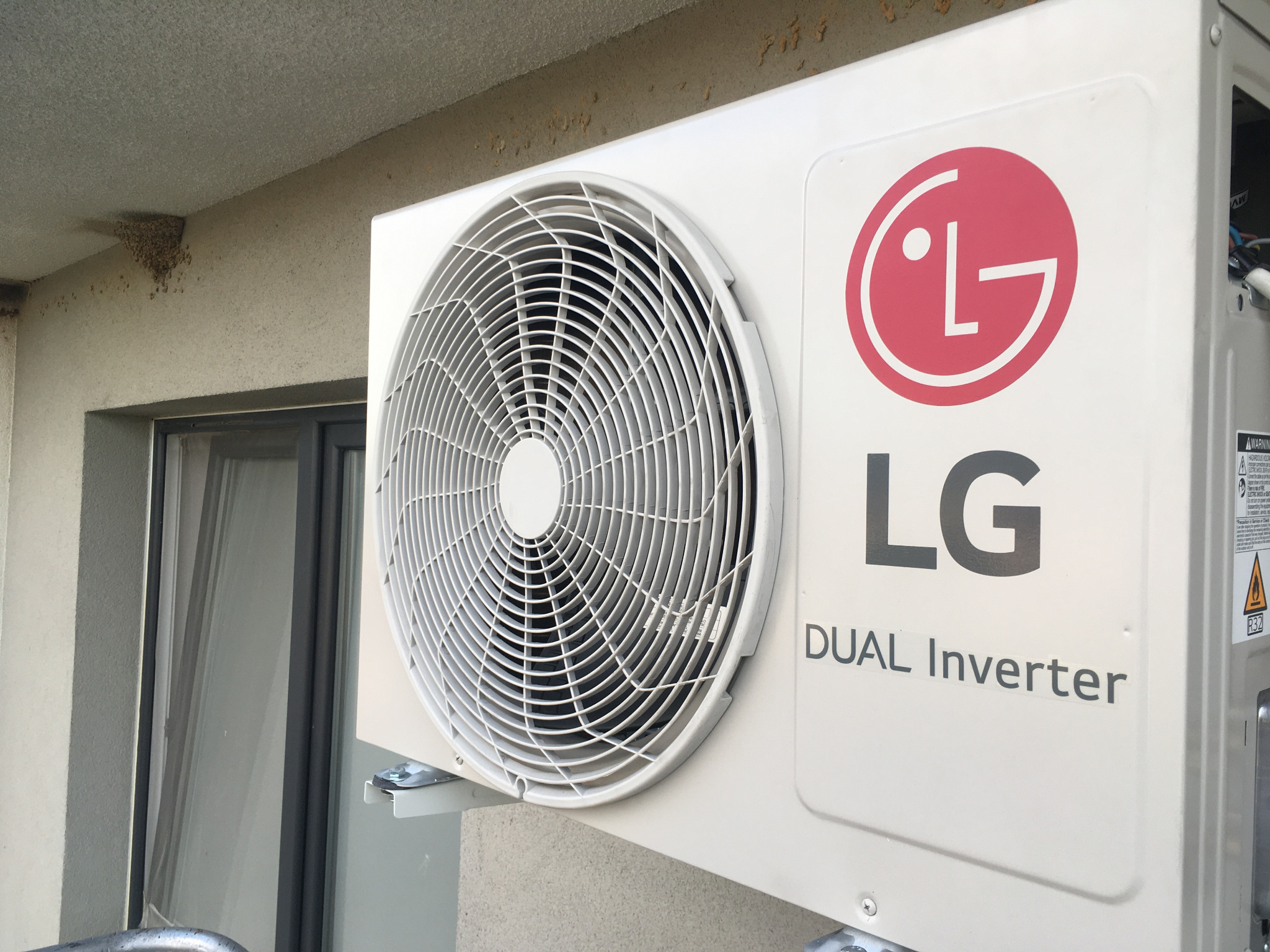 Zewnętrzna jednostka klimatyzacji LG Dual Inverter zamontowana na ścianie budynku, widoczne gniazdo jaskółcze pod okapem.