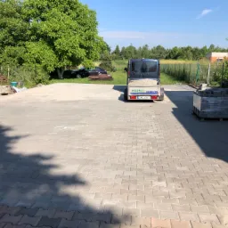 Rozległy plac wyłożony szarą kostką brukową, w tle zaparkowany samochód i przyczepa z piaskiem, paleta z kostką po prawej stronie, cień drzewa na kostce.