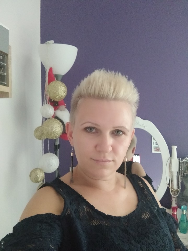 Portret kobiety z krótkimi blond włosami, w czarnej ażurowej bluzce z odkrytymi ramionami i długimi kolczykami na tle fioletowej ściany i białych mebli z dekoracjami.