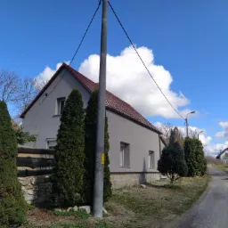 Szary dom jednorodzinny z czerwoną dachówką, widziany z poziomu ulicy, z elementami ogrodzenia i zieleni na pierwszym planie, na tle błękitnego nieba z białymi chmurami.