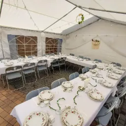 Uroczysty stół, nakryty białą zastawą i ozdobiony girlandami, ustawiony wewnątrz białego namiotu z dekoracjami wiszącymi pod sufitem, przygotowany na przyjęcie.