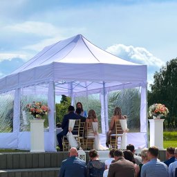 DekoPlener Wypożyczalnia Namiotów i Dekoracji - Ślub w plenerze pod białym namiotem z przeźroczystymi ścianami. Goście siedzą na ławkach, a para młoda na złotych krzesłach. Na podwyższeniu dekoracje kwiatowe.