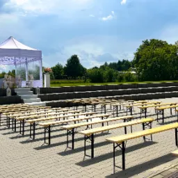 Plenerowa aranżacja ślubna: biały namiot z dekoracjami kwiatowymi na podwyższeniu, rzędy drewnianych ławek dla gości, tło z zieleni i stóg siana.