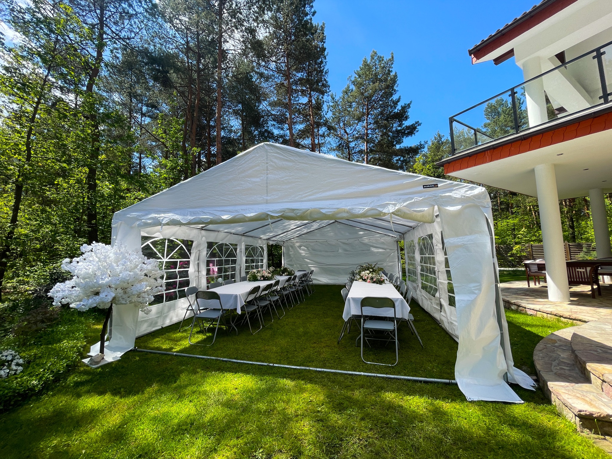 Biały namiot plenerowy na trawie, ustawiony na przyjęcie, z dekoracjami kwiatowymi na stołach, w tle las i budynek z balkonem.