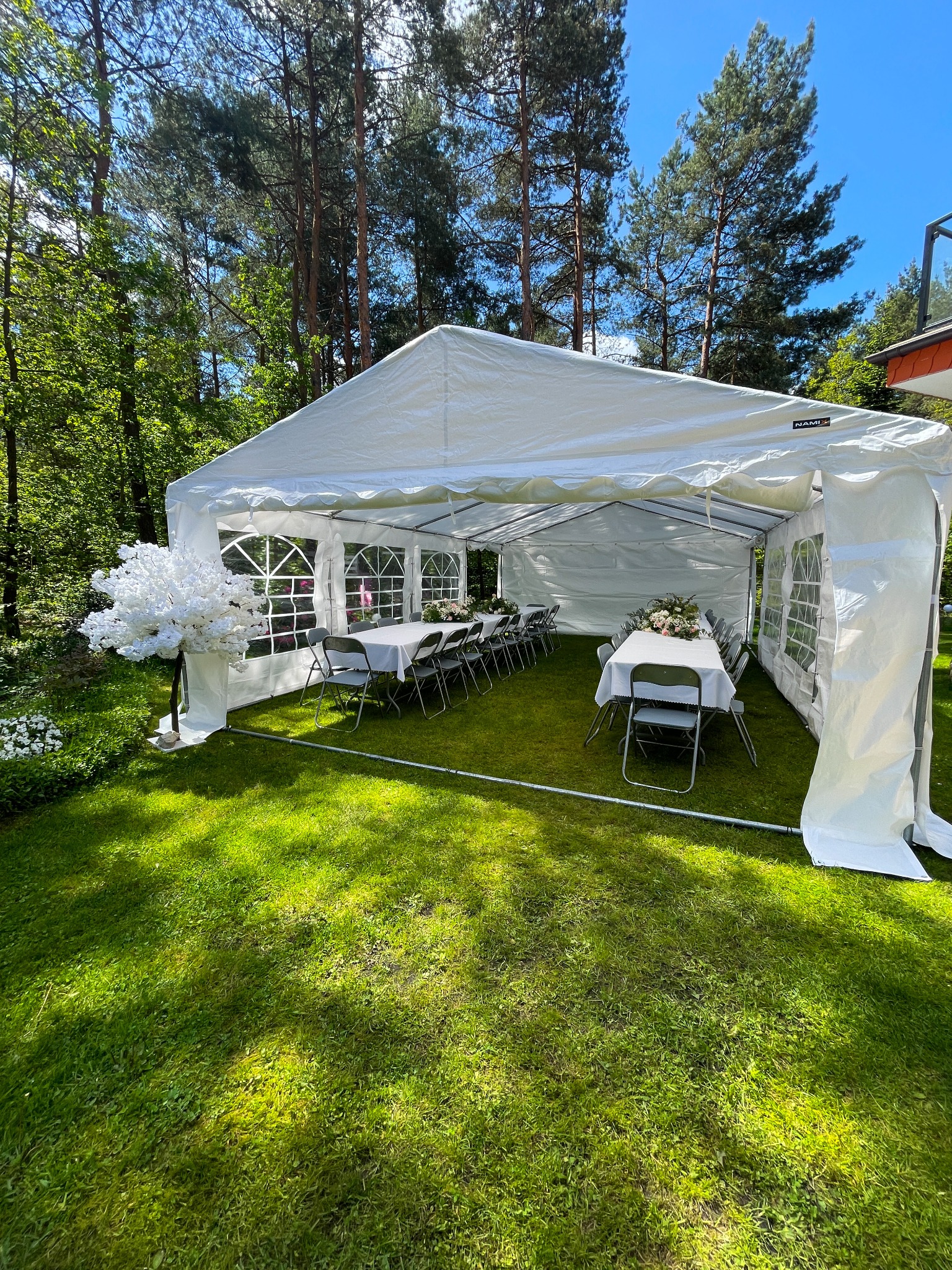 Biały namiot imprezowy na trawie, ustawiony na przyjęcie. Wnętrze z nakrytymi stołami i krzesłami. Dekoracja kwiatowa i sztuczne drzewko.