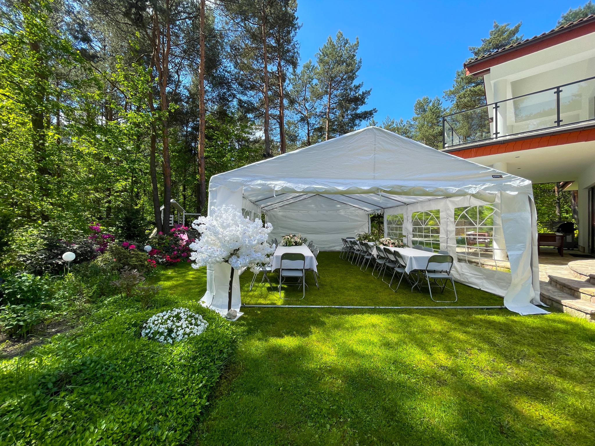 Biały namiot imprezowy na zielonym trawniku, przygotowany na przyjęcie z ustawionymi stołami i krzesłami, dekoracjami kwiatowymi. W tle dom i drzewa.