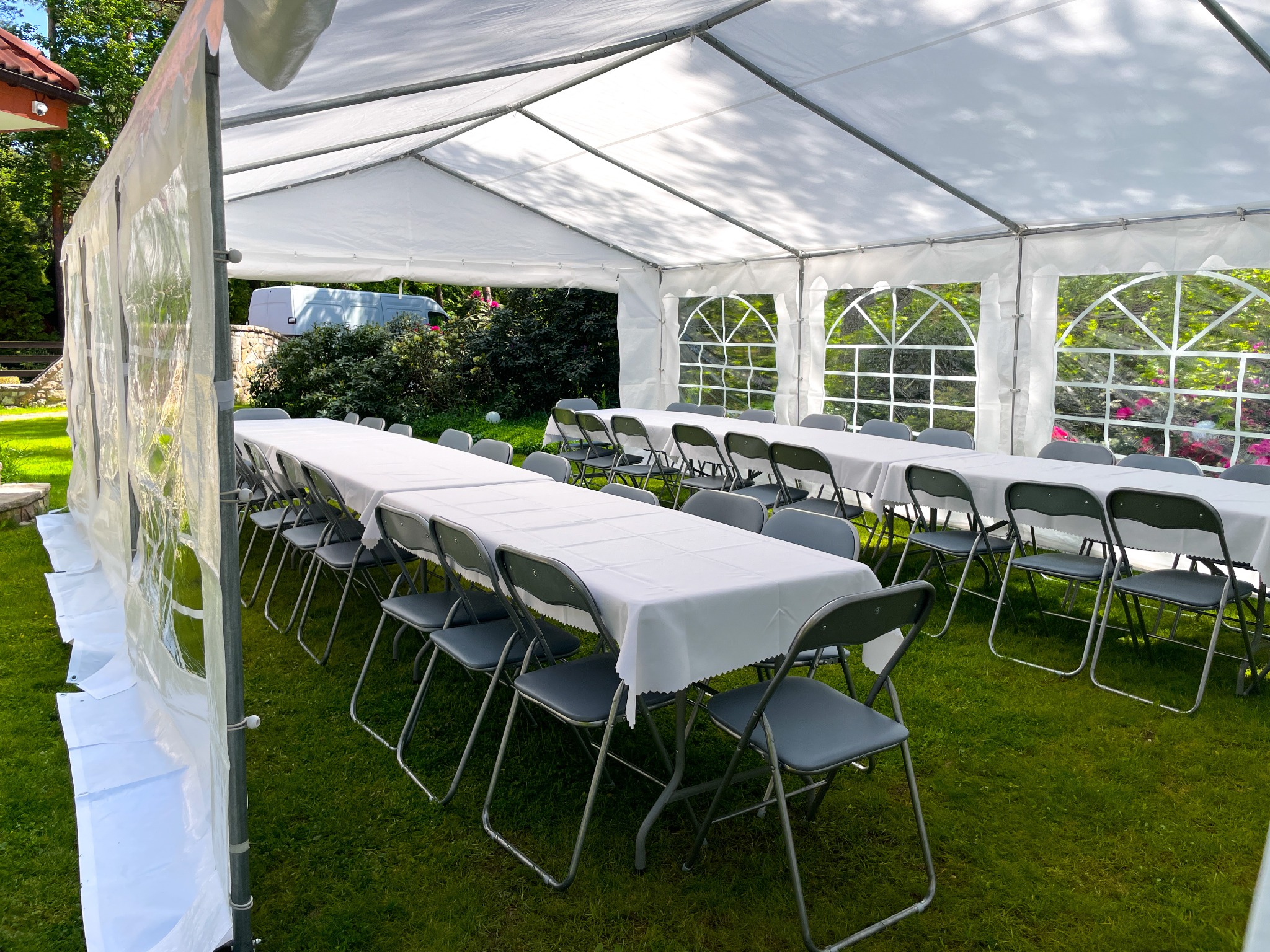 Biały namiot bankietowy na trawie, przygotowany na imprezę. Widoczne stoły z białymi obrusami i krzesła. W tle zieleń drzew i budynek. Całość oświetlona naturalnym światłem.