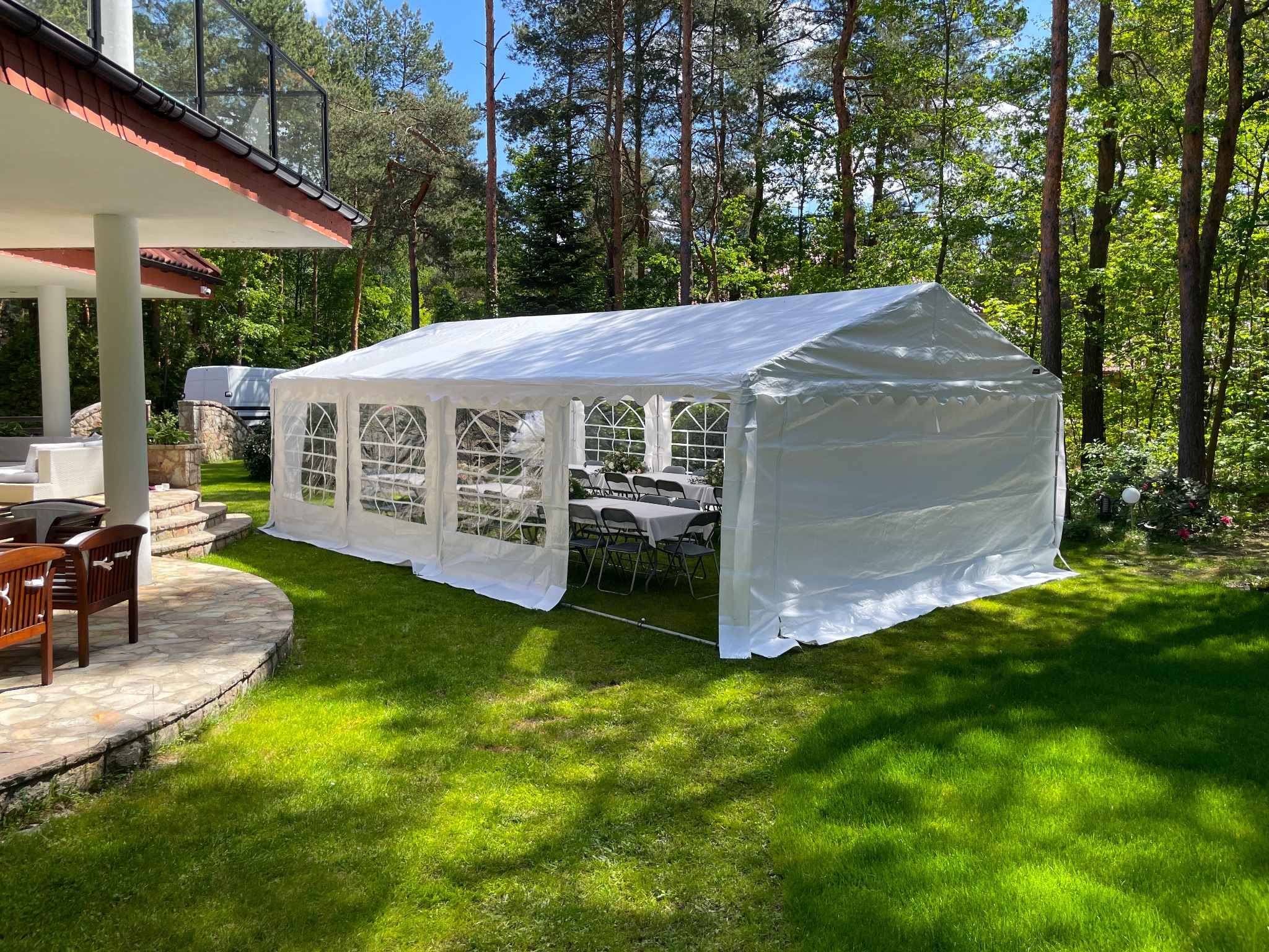 Biały namiot bankietowy z oknami, ustawiony na trawniku obok tarasu domu, w tle drzewa. W namiocie widoczne stoły i krzesła.