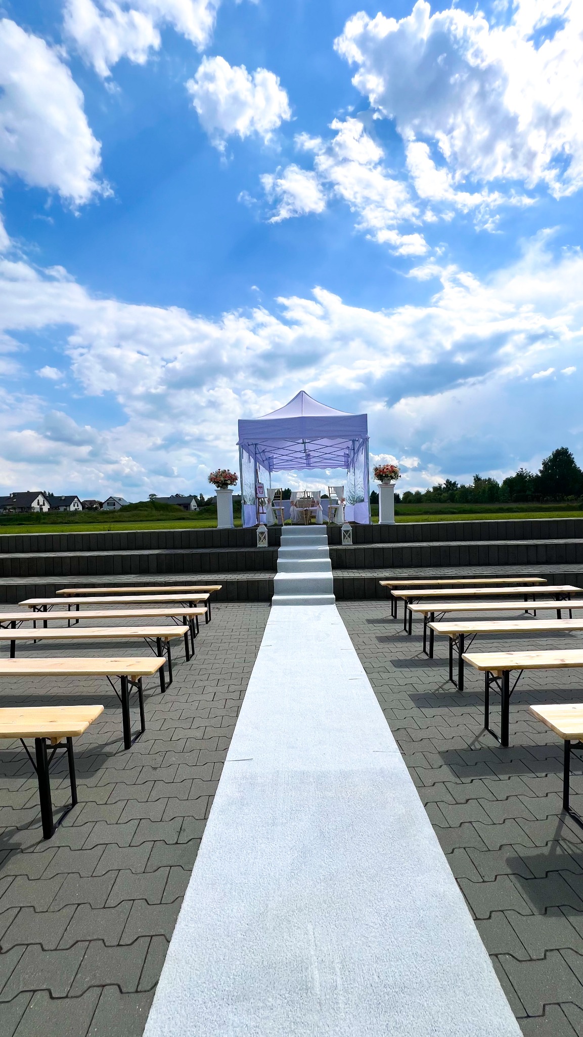 Plenerowa aranżacja ślubna: biały dywan prowadzi do namiotu z kwiatami, otoczonego drewnianymi ławkami na tle błękitnego nieba z chmurami.