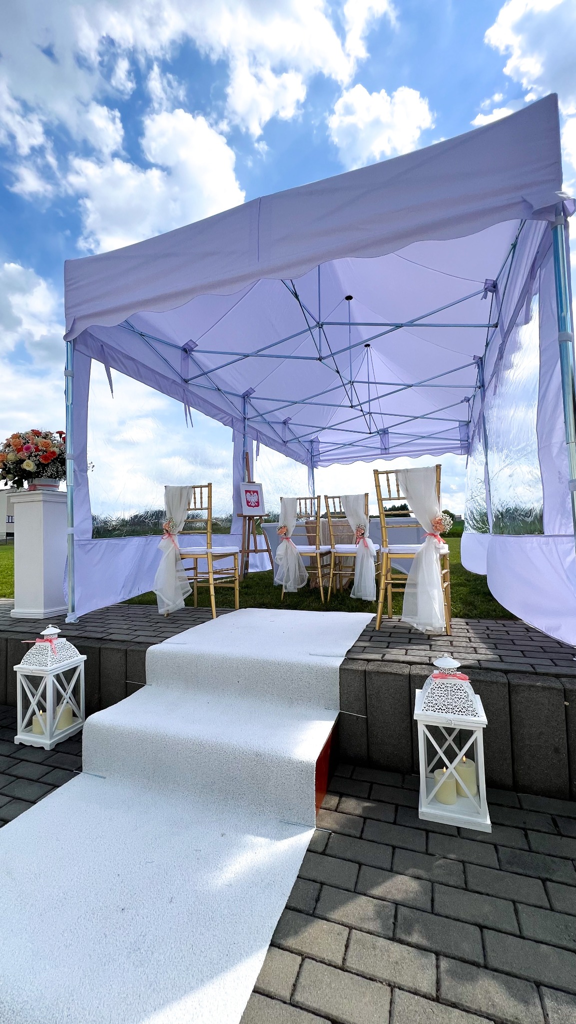 Biały namiot plenerowy z dekoracjami ślubnymi: krzesła, kwiaty, lampiony. Ustawiony na zewnątrz, w tle błękitne niebo z chmurami. Dekoracja w stylu rustykalnym.