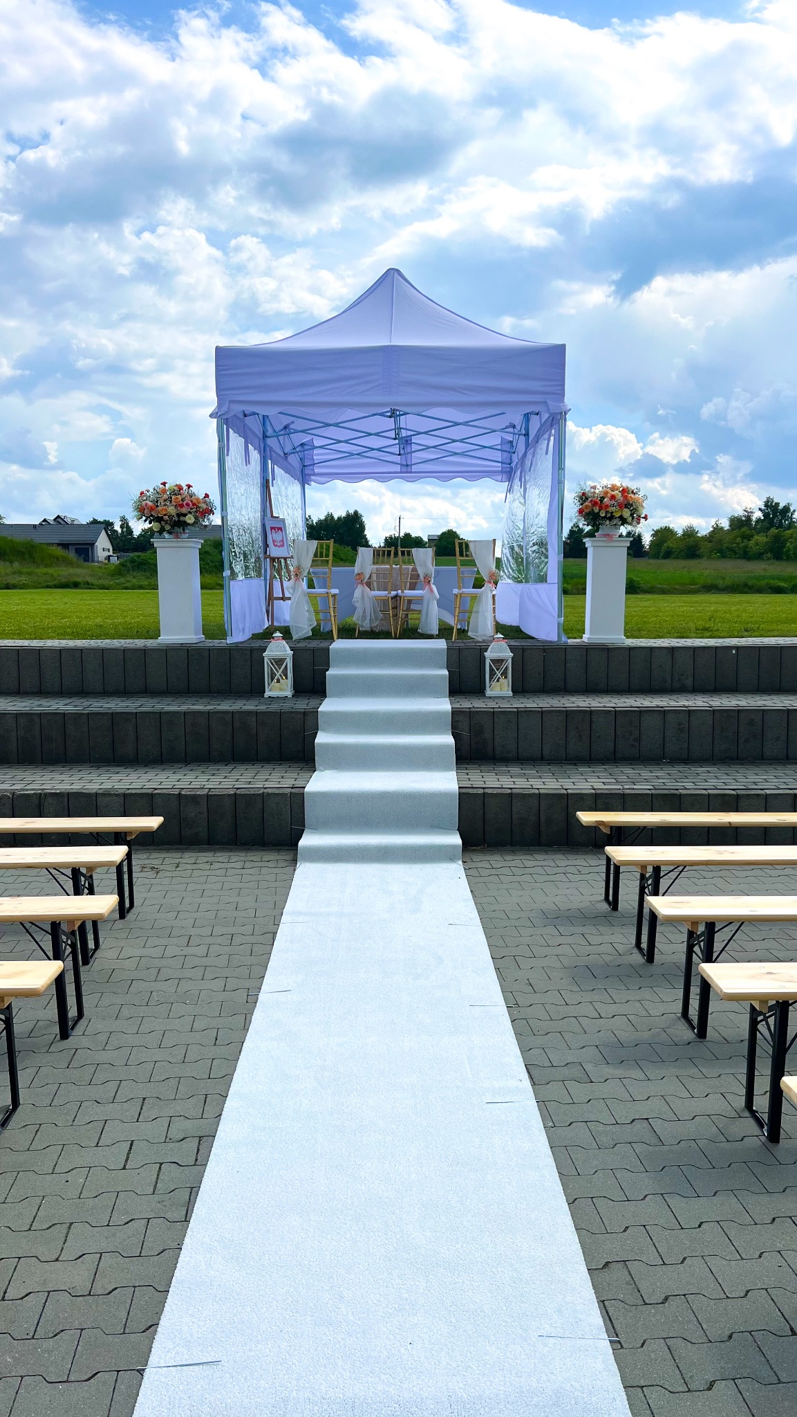 Biały namiot plenerowy z dekoracją kwiatową i ścieżką na schodach, przygotowany na ślub lub uroczystość, w tle błękitne niebo.