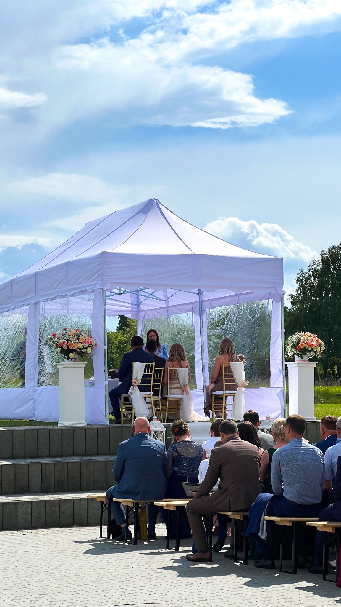 Ślub w plenerze pod białym namiotem z przeźroczystymi ścianami. Goście siedzą na ławkach, a para młoda na złotych krzesłach. Na podwyższeniu dekoracje kwiatowe.