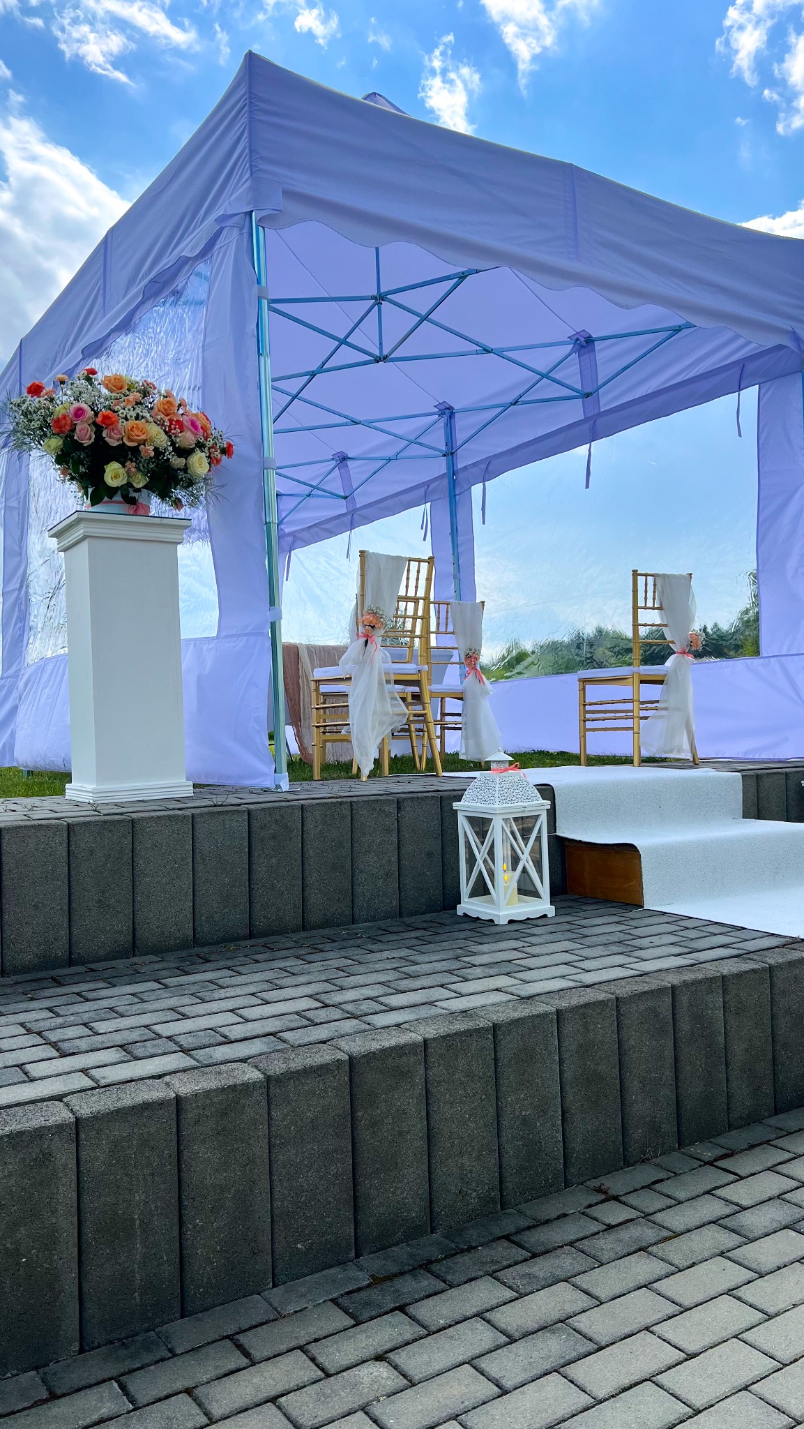 Namiot ślubny w plenerze: biały, z dekoracjami kwiatowymi i lampionem, ustawiony na podwyższeniu z kostki brukowej. Krzesła udekorowane wstążkami i tkaniną.