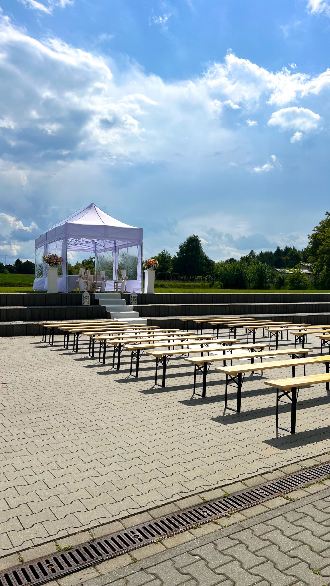 Biały namiot plenerowy z dekoracjami kwiatowymi, ustawiony na podwyższeniu z rzędami drewnianych ławek. Błękitne niebo z chmurami w tle.