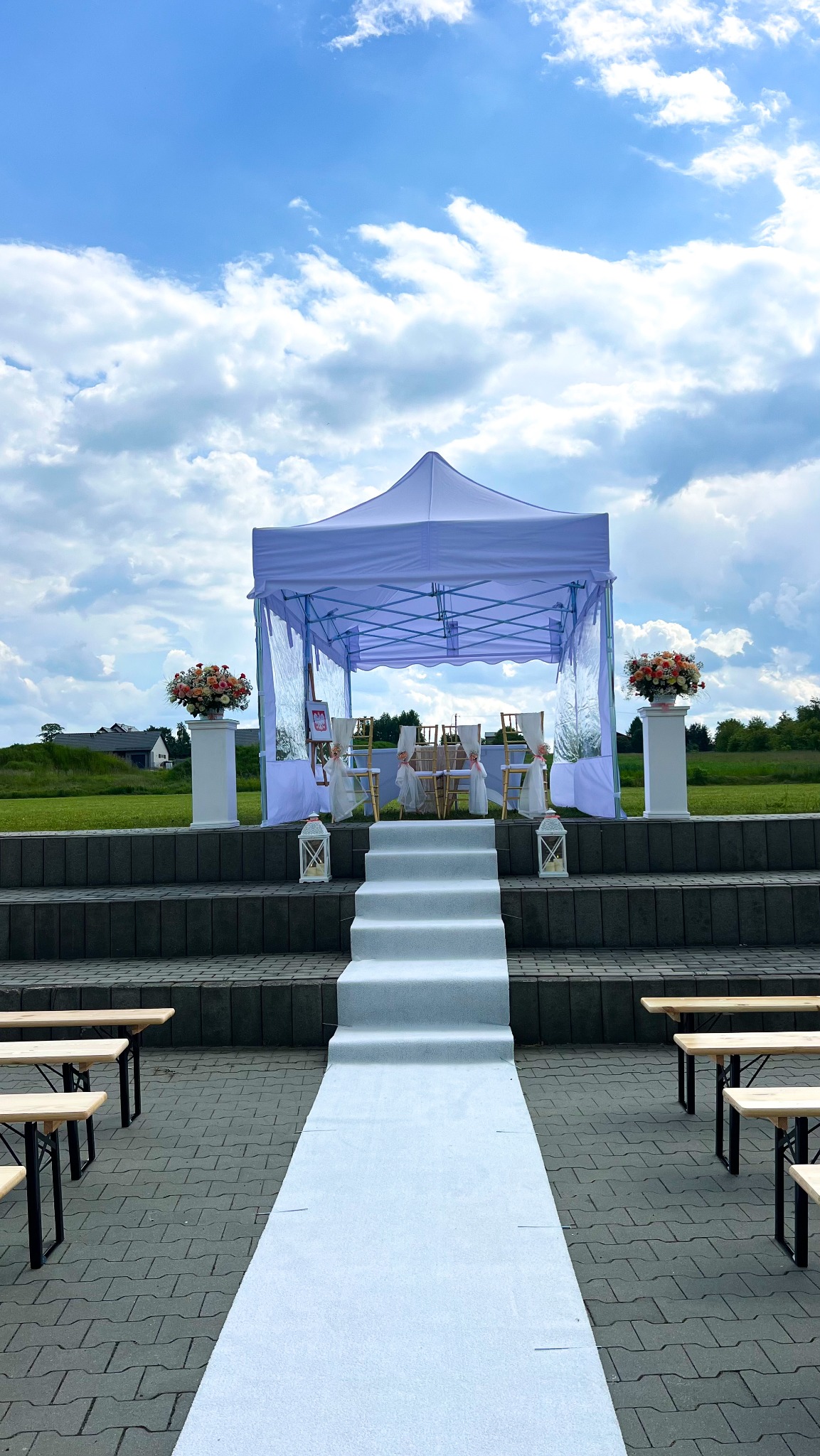 Biały namiot plenerowy z dekoracjami kwiatowymi i ścieżką, przygotowany na ceremonię ślubną na zewnątrz. Widok na zielone pola i błękitne niebo z chmurami.