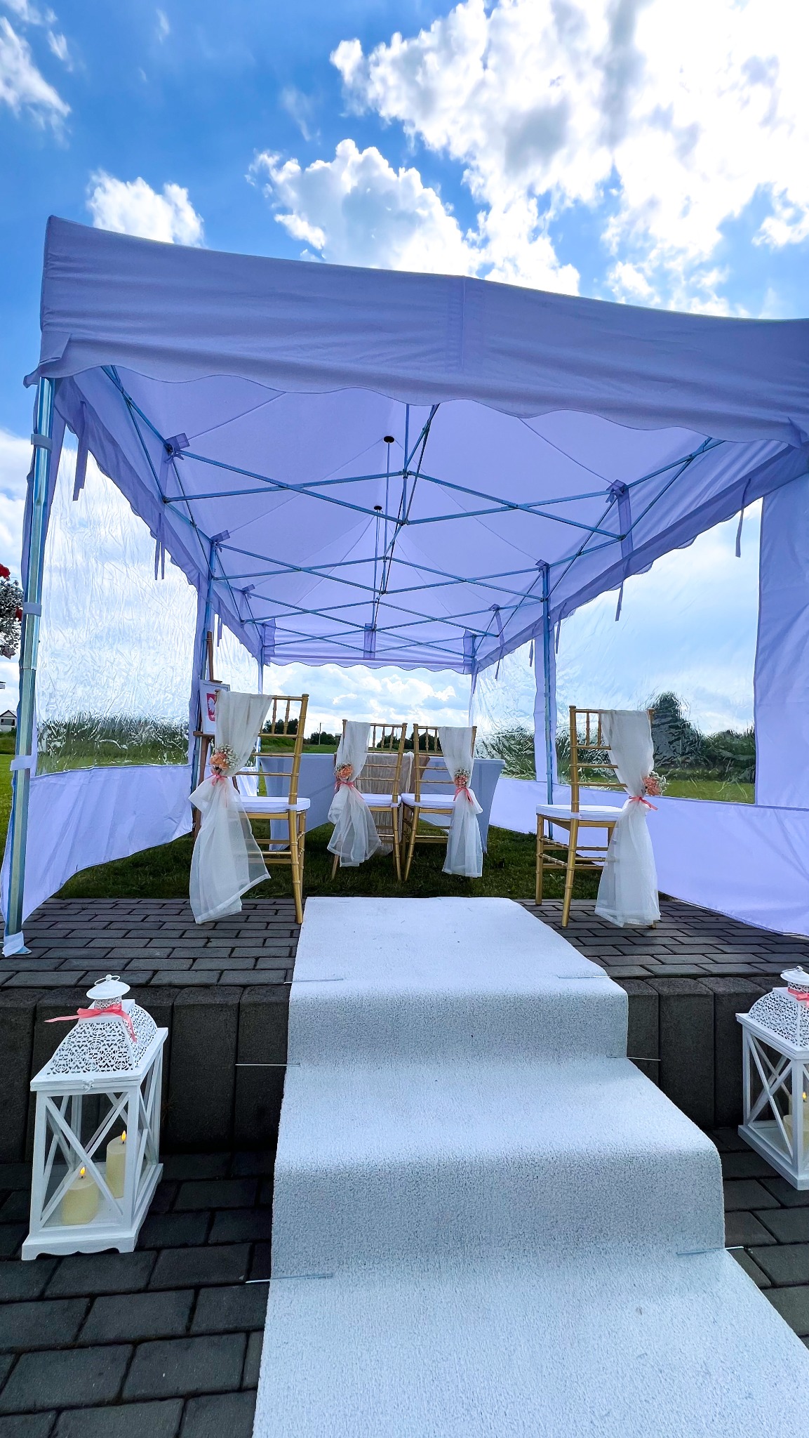 Biały namiot z transparentnymi ścianami, dekorowane krzesła, biały dywan i lampiony tworzą elegancką scenerię plenerową na ślub lub inną uroczystość.