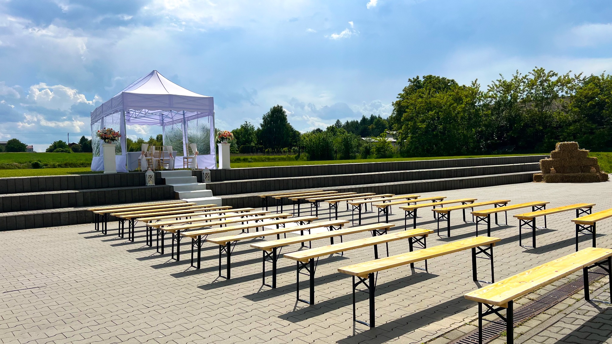 Plenerowa aranżacja ślubna: biały namiot z dekoracjami kwiatowymi na podwyższeniu, rzędy drewnianych ławek dla gości, tło z zieleni i stóg siana.