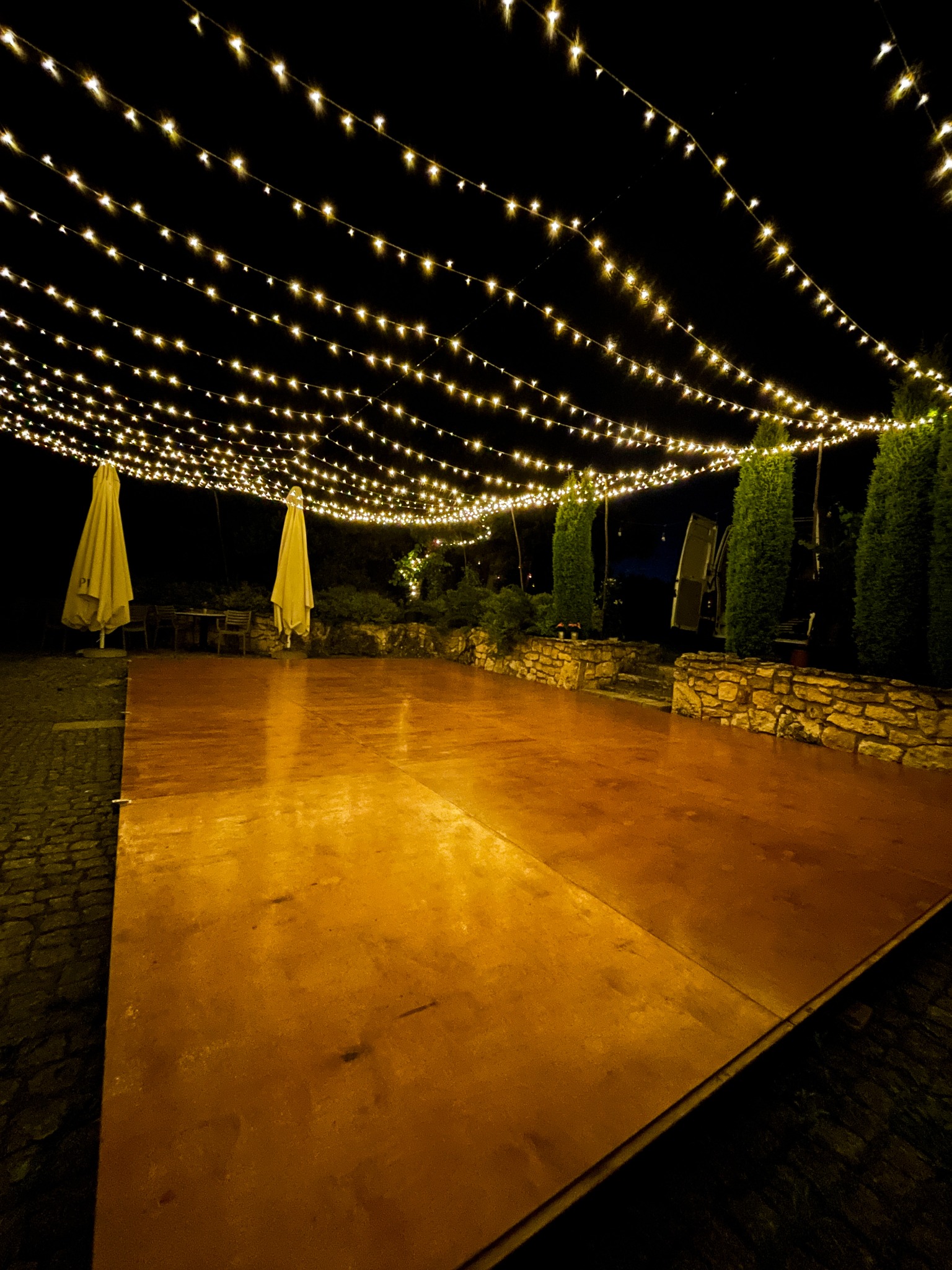 Parkiet taneczny 5x8 m oświetlony fairy lights, tworzący efekt gwieździstego nieba. Dwa złożone parasole w tle, otoczenie zieleni i kamienny murek.