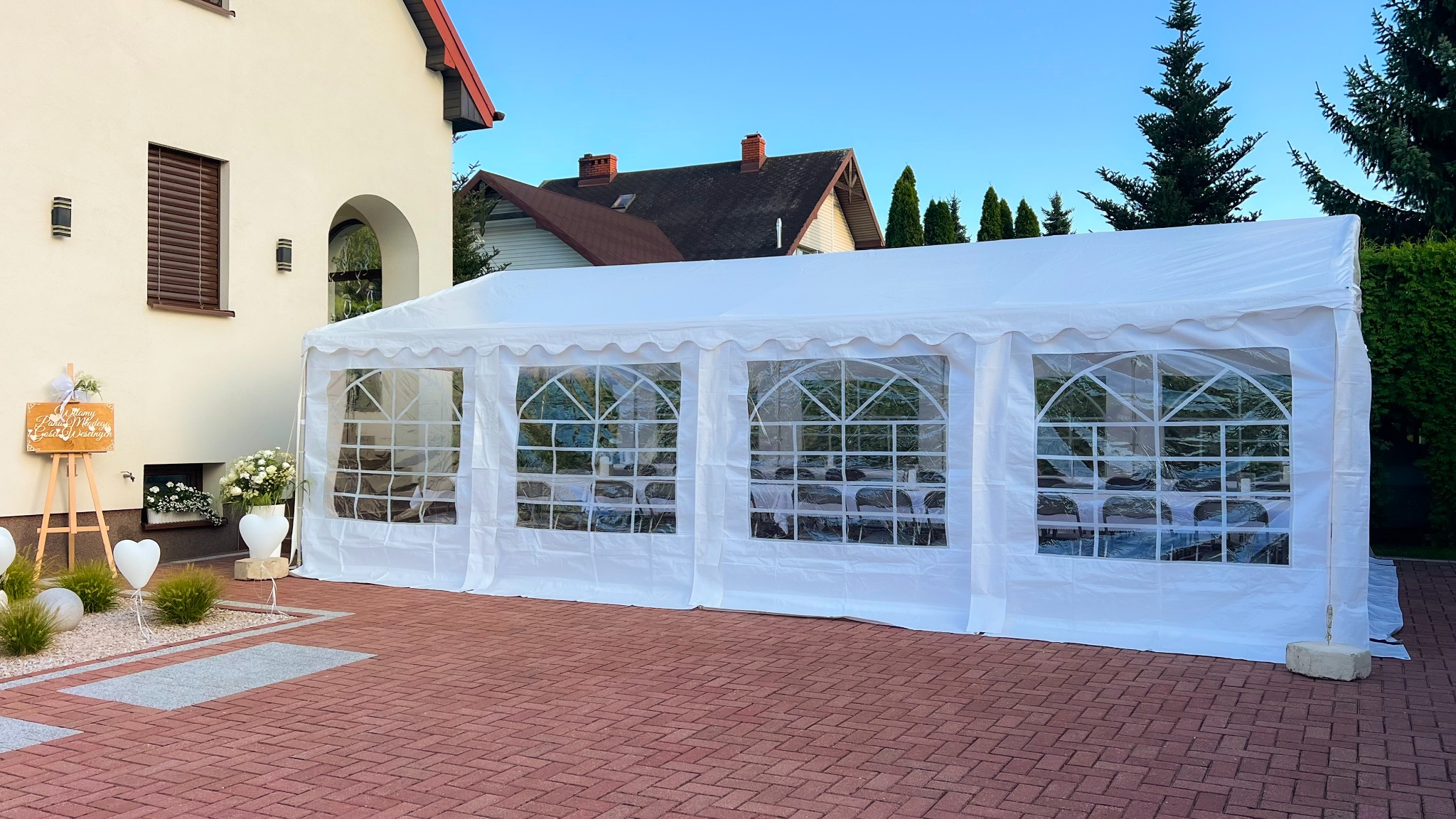 Biały namiot z oknami na ceglanym podwórku, dekoracje ślubne, tablica powitalna, dom w tle, błękitne niebo. Garden party w plenerze.