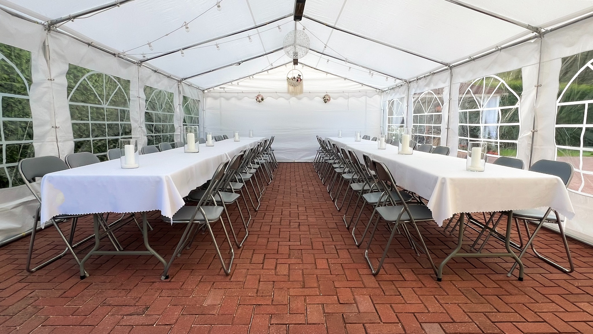Biały namiot imprezowy z rzędami stołów nakrytych białymi obrusami i szarymi krzesłami, dekoracje wiszące, ceglana podłoga, widok z perspektywy środka namiotu.