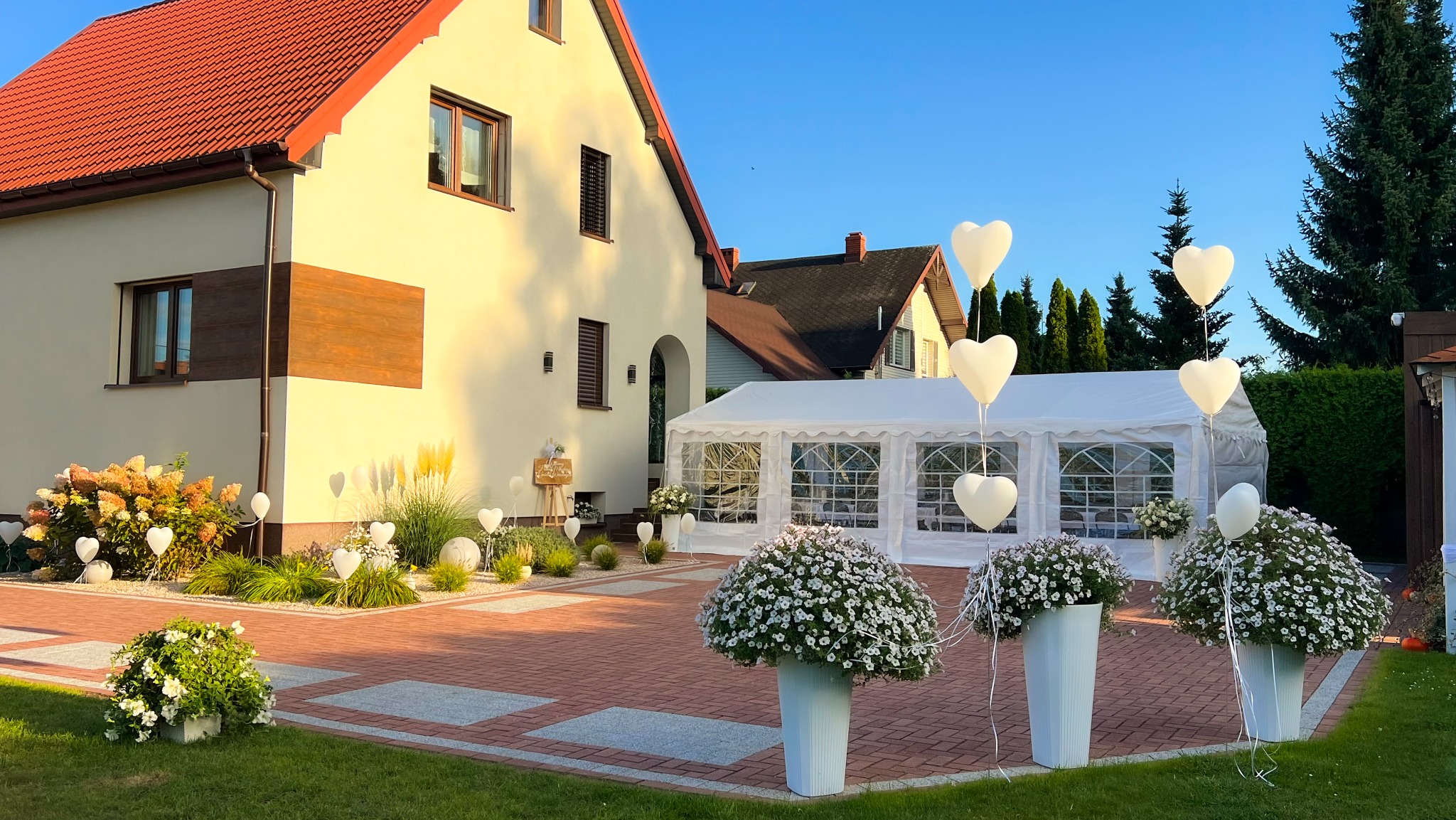 Biały namiot imprezowy z dekoracjami (balony w kształcie serc, kwiaty) na podwórku domu z czerwoną dachówką, w słoneczny dzień. Garden party w plenerze.