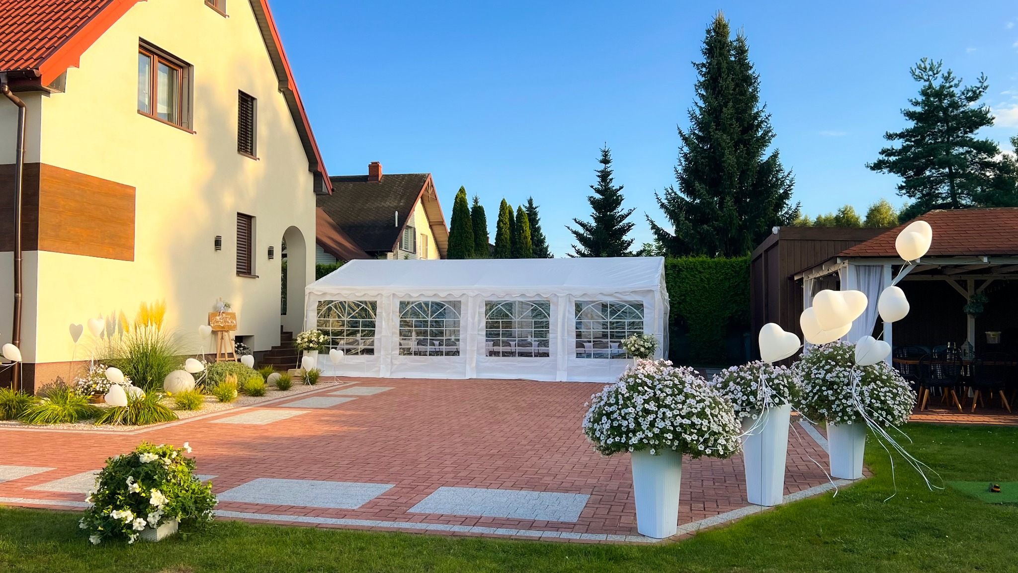 Biały namiot imprezowy z oknami na podwórku, dekoracje kwiatowe w donicach i białe balony w kształcie serc, dom w tle, słoneczny dzień.