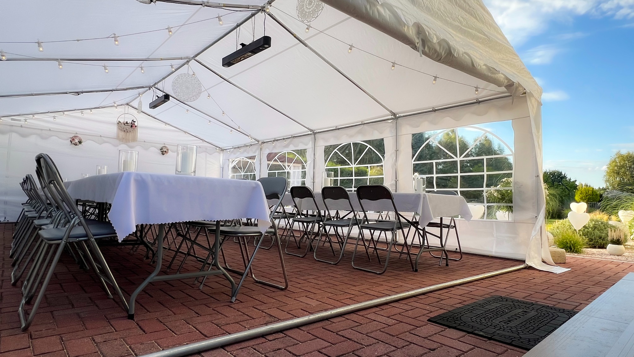 Ustawiony namiot imprezowy z oknami, stołami nakrytymi białymi obrusami i krzesłami. Dekoracje wiszące pod sufitem, ceglana podłoga i widok na zewnątrz.