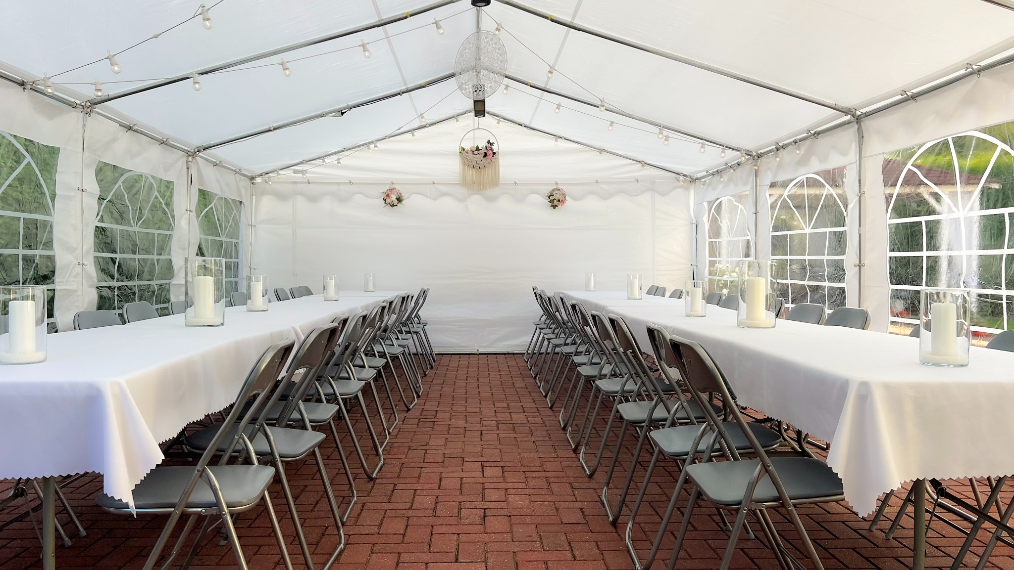 Uroczysty namiot z białymi obrusami, świecami w szklanych wazonach i dekoracjami kwiatowymi, ustawiony na ceglanym podłożu, oświetlony girlandami żarówek.