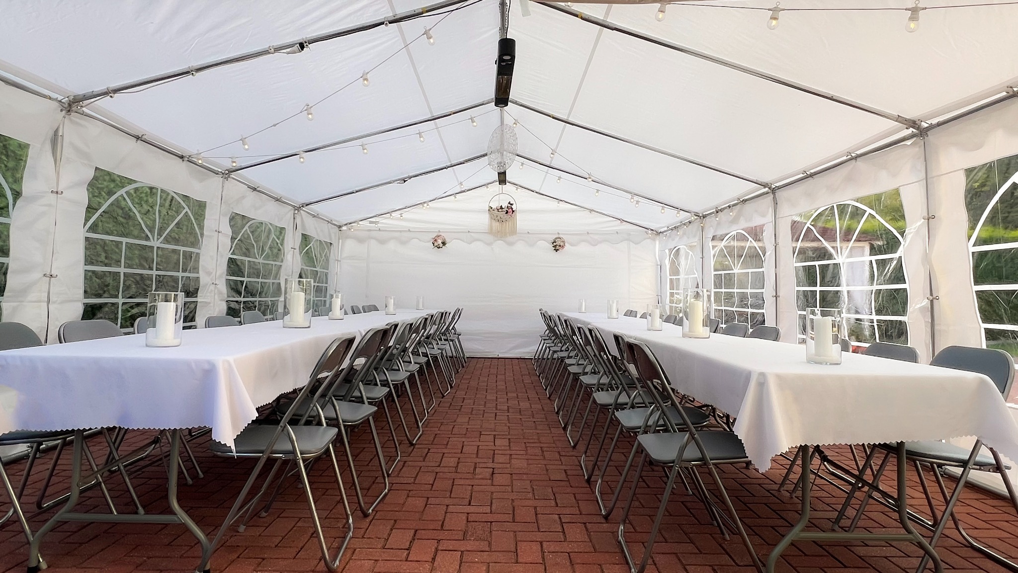 Ustawiony namiot imprezowy z długimi stołami nakrytymi białymi obrusami, szarymi krzesłami i dekoracjami, oświetlony girlandami żarówek.