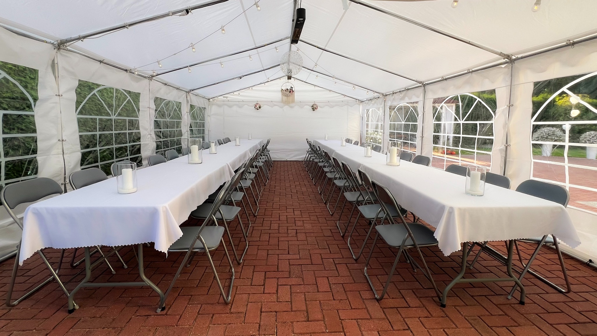 Ustawiony namiot imprezowy z dwoma długimi stołami nakrytymi białymi obrusami, szarymi krzesłami i świecami w szklanych lampionach. Czerwona ceglana podłoga, dekoracje.