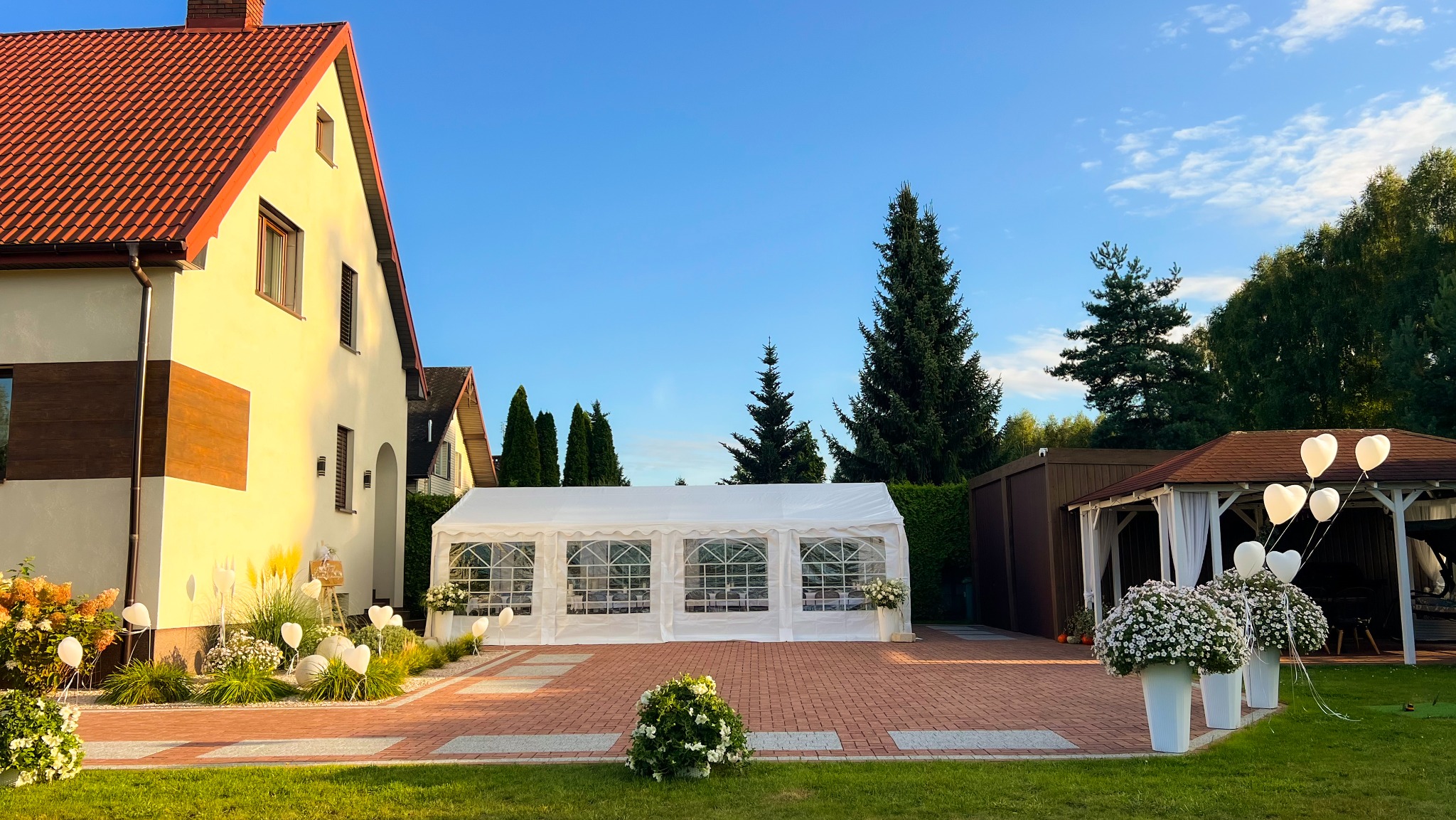 Biały namiot imprezowy na ceglanym podwórku, dekorowany kwiatami i balonami w kształcie serc, ustawiony obok domu z czerwonym dachem, w słoneczny dzień.