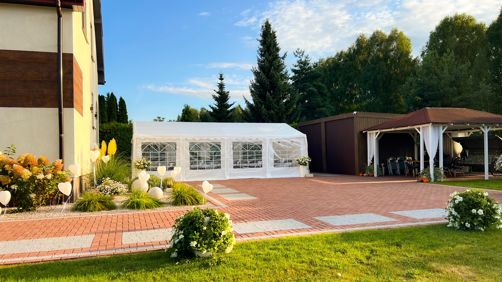 Biały namiot imprezowy z dekoracjami na ceglanym podwórku, obok budynku i altany. Udekorowany ogród z kwiatami i balonami w kształcie serc.