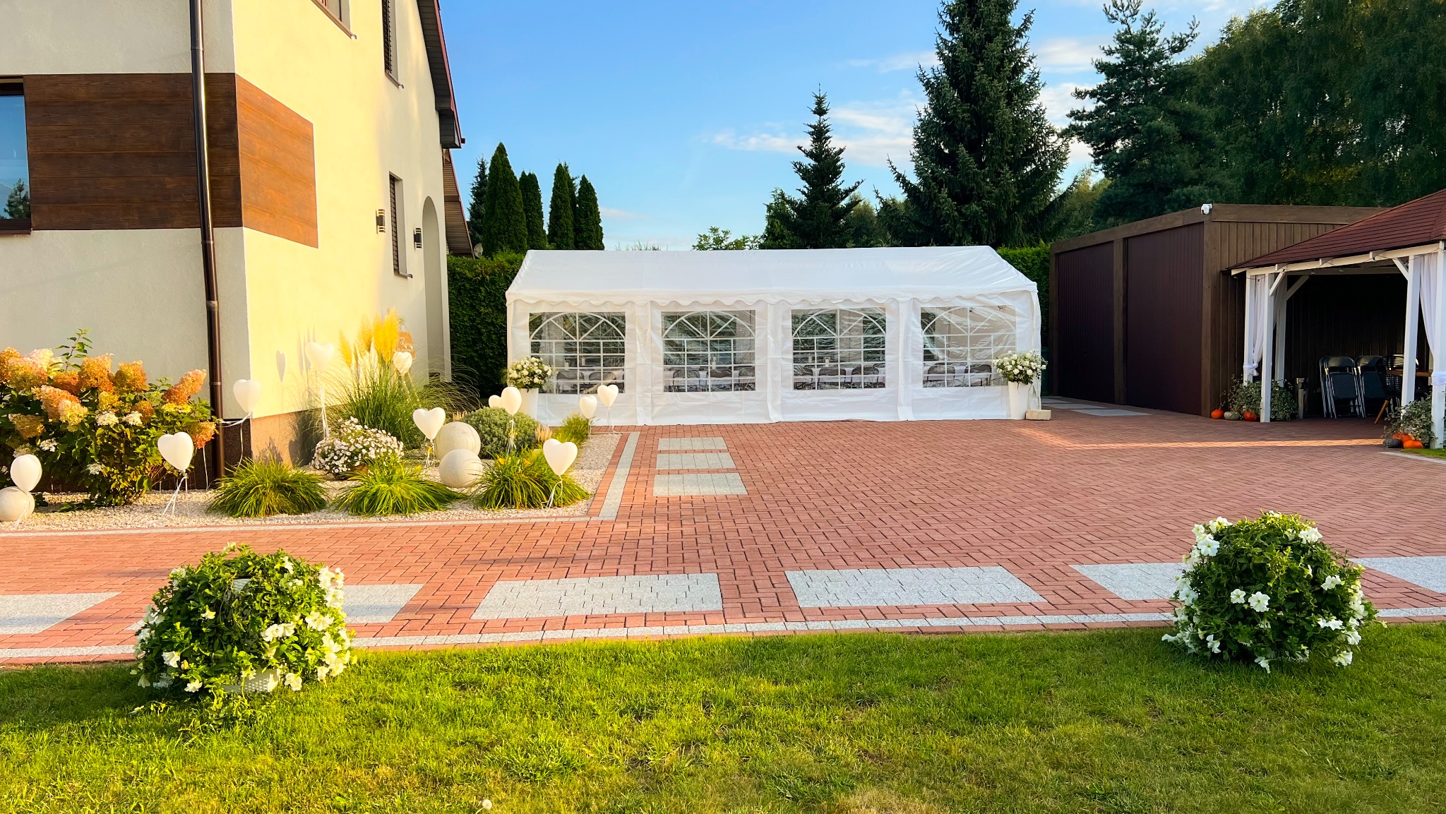 Biały namiot imprezowy z dekoracjami kwiatowymi i balonami w kształcie serc, ustawiony na podwórku z kostki brukowej, idealny na garden party.