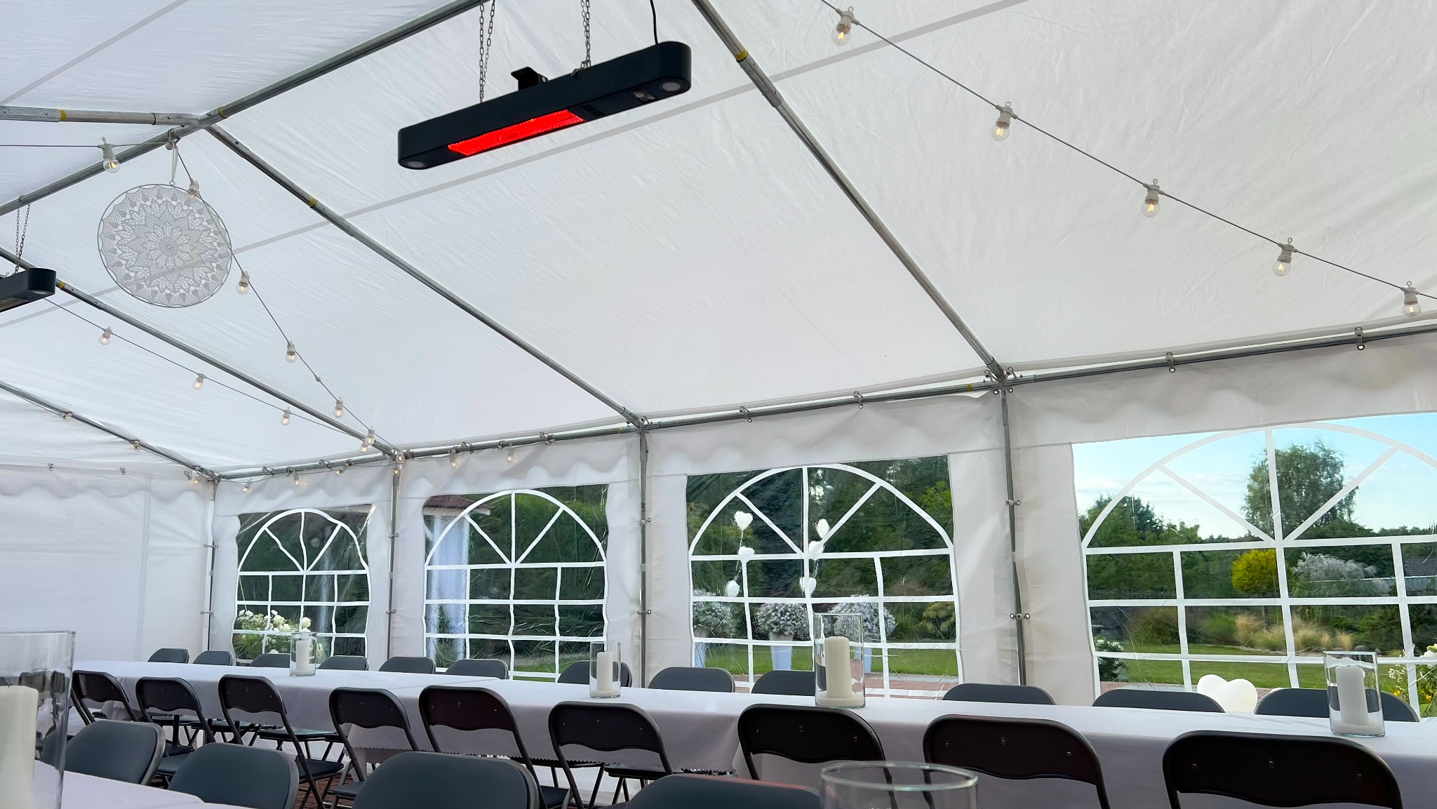 Biały namiot imprezowy z oknami i dekoracjami w Częstochowie. Długi stół z krzesłami, oświetlenie girlandami i grzejnikami. Widok na zewnątrz przez okna.