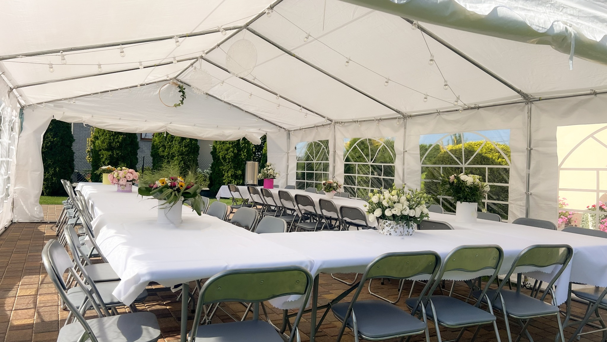 Biały namiot imprezowy z dekoracjami kwiatowymi, stołami nakrytymi białymi obrusami i szarymi krzesłami. Widok na zewnątrz przez okna namiotu.