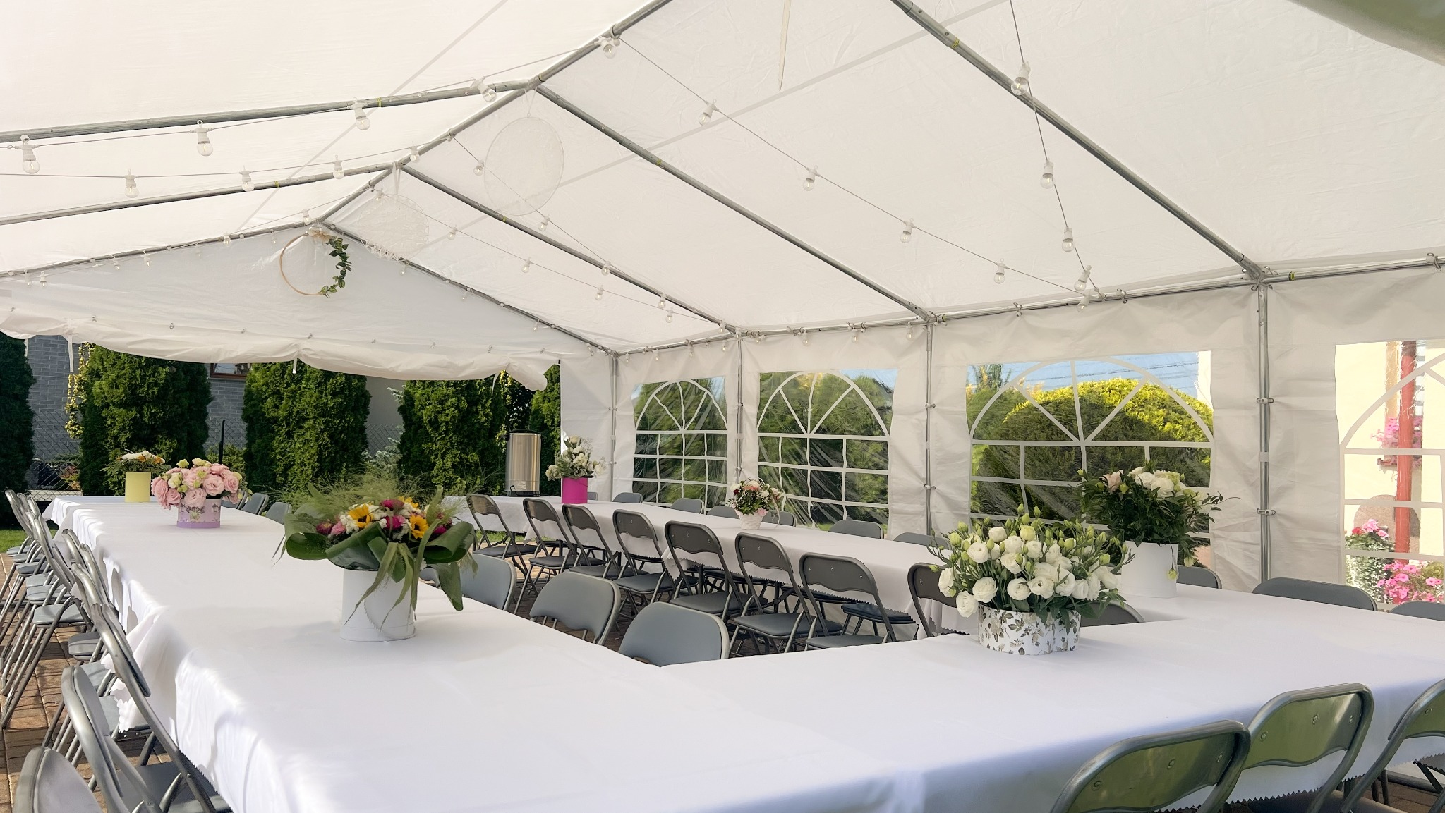 Biały namiot imprezowy z oknami, oświetleniem i dekoracjami kwiatowymi. Stoły nakryte białymi obrusami, ustawione w podkowę, otoczone szarymi krzesłami.