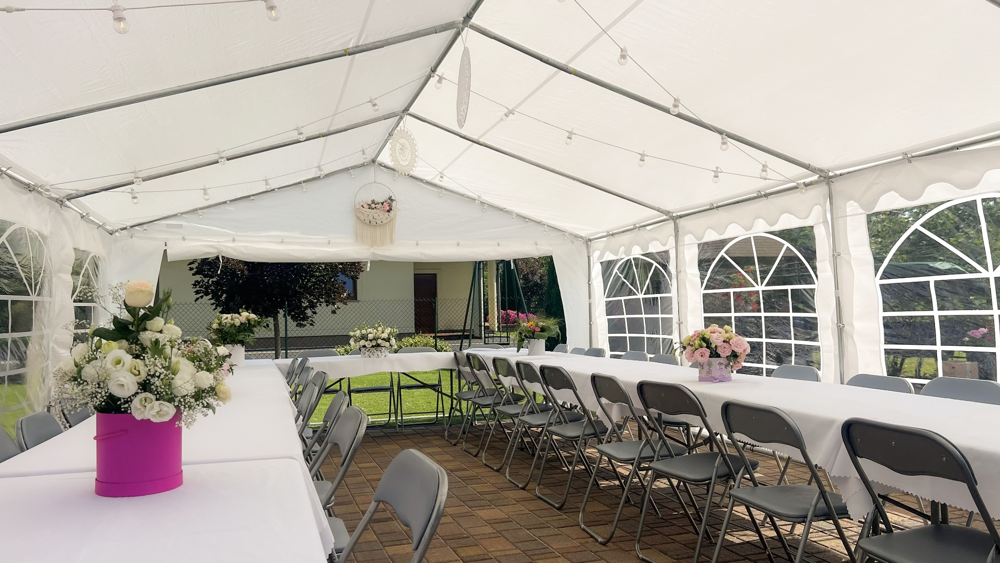 Biały namiot imprezowy z dekoracjami: stoły z obrusami, krzesła, oświetlenie girlandami, kwiatowe kompozycje. Widok na zewnątrz przez okna namiotu.