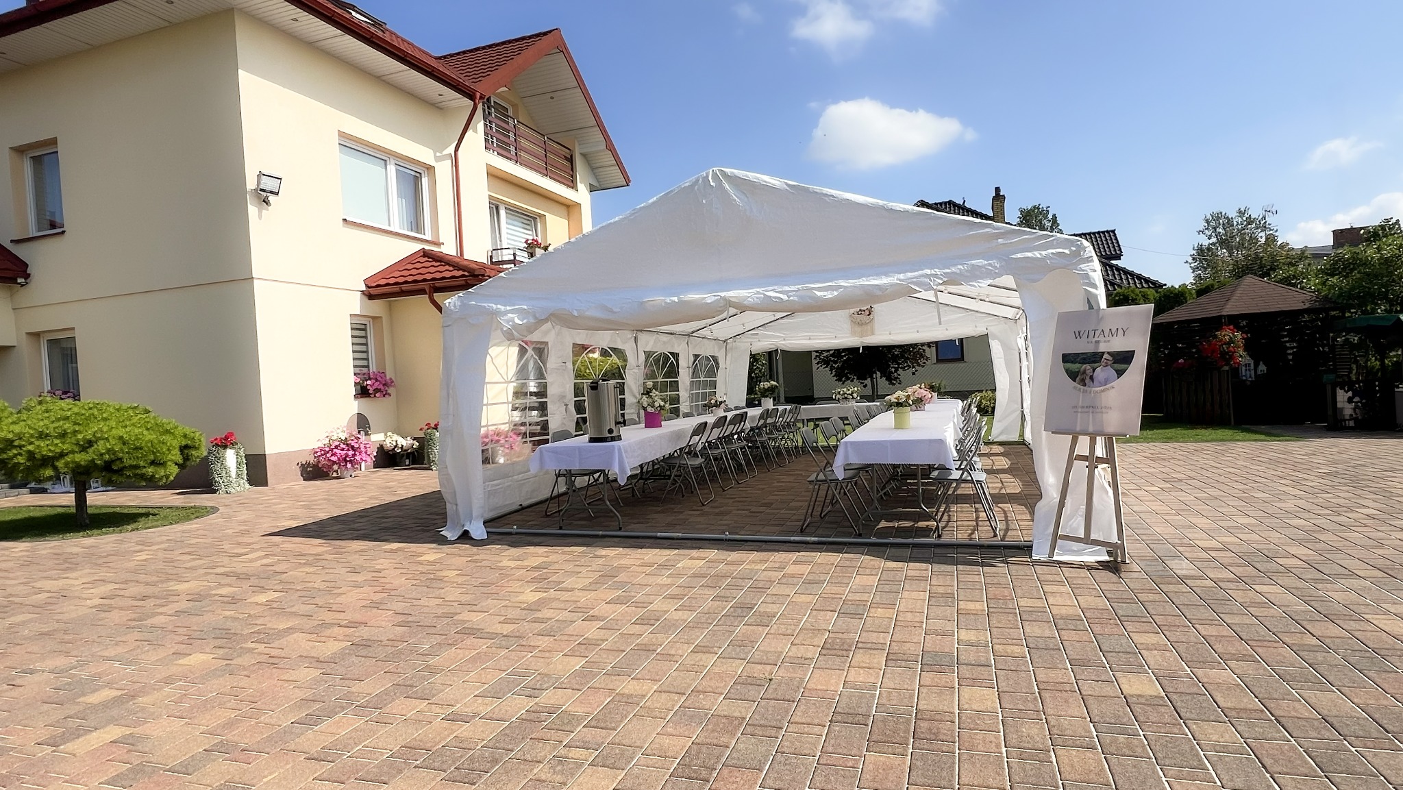 Biały namiot imprezowy z wyposażeniem (stoły, krzesła, dekoracje) ustawiony na zewnątrz, przed domem. Widoczny baner powitalny na sztaludze.
