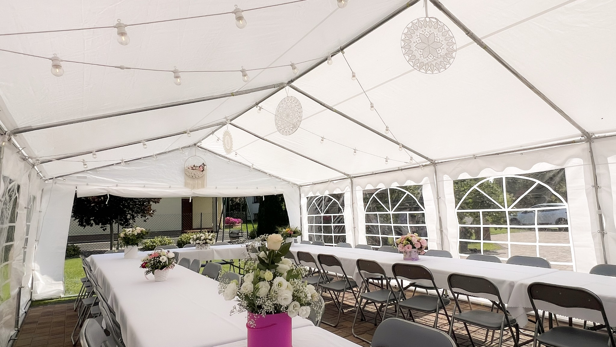 Udekorowany namiot imprezowy z oświetleniem, stołami i krzesłami, przygotowany na przyjęcie. Widoczne dekoracje kwiatowe i ozdobne lampiony.