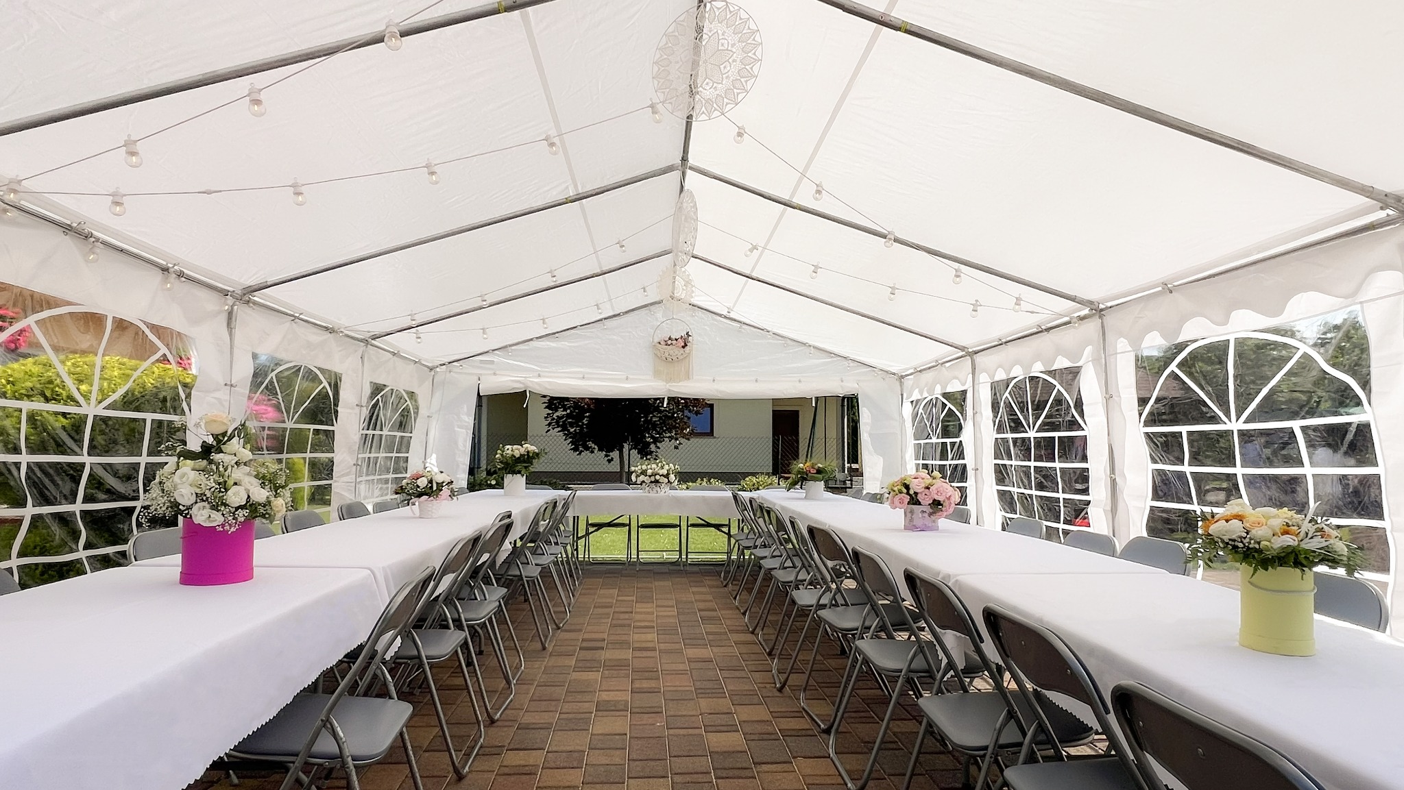 Udekorowany namiot imprezowy z oświetleniem, stołami nakrytymi białymi obrusami i krzesłami. Kompozycje kwiatowe w pastelowych kolorach zdobią stoły. Widok z wnętrza namiotu.