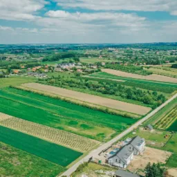 Szeroki kadr z lotu ptaka ukazuje mozaikę pól uprawnych w odcieniach zieleni i beżu, z nowo wybudowanymi domami jednorodzinnymi z szarymi dachami w dolnej części kadru, przeciętych wąską, asfaltową...