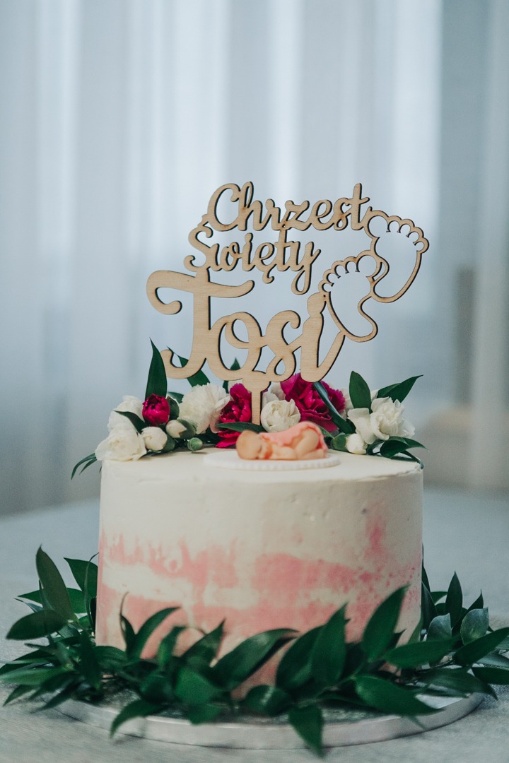 Tort na chrzest z drewnianym topperem 'Chrzest Święty Tosi', dekorowany kwiatami i figurką śpiącego niemowlęcia.